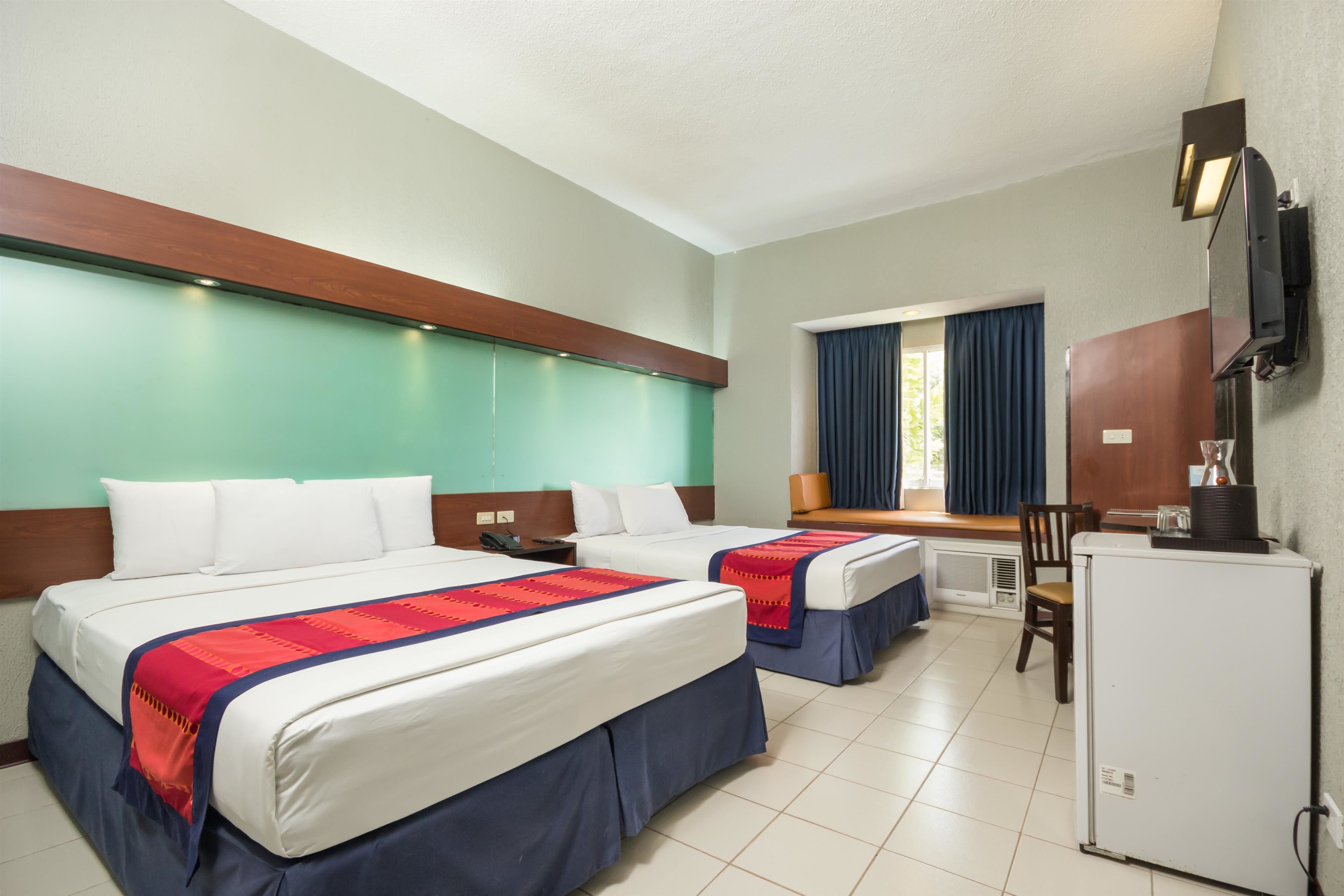 A hotel room with two beds, a desk, a chair, a refrigerator, and a television mounted on the wall.