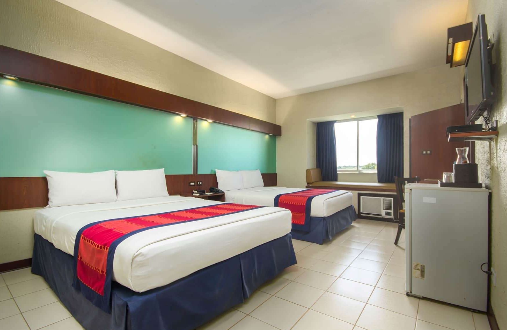 Habitación de hotel con dos camas, una kitchenette pequeña, un televisor y una ventana con cortinas.
