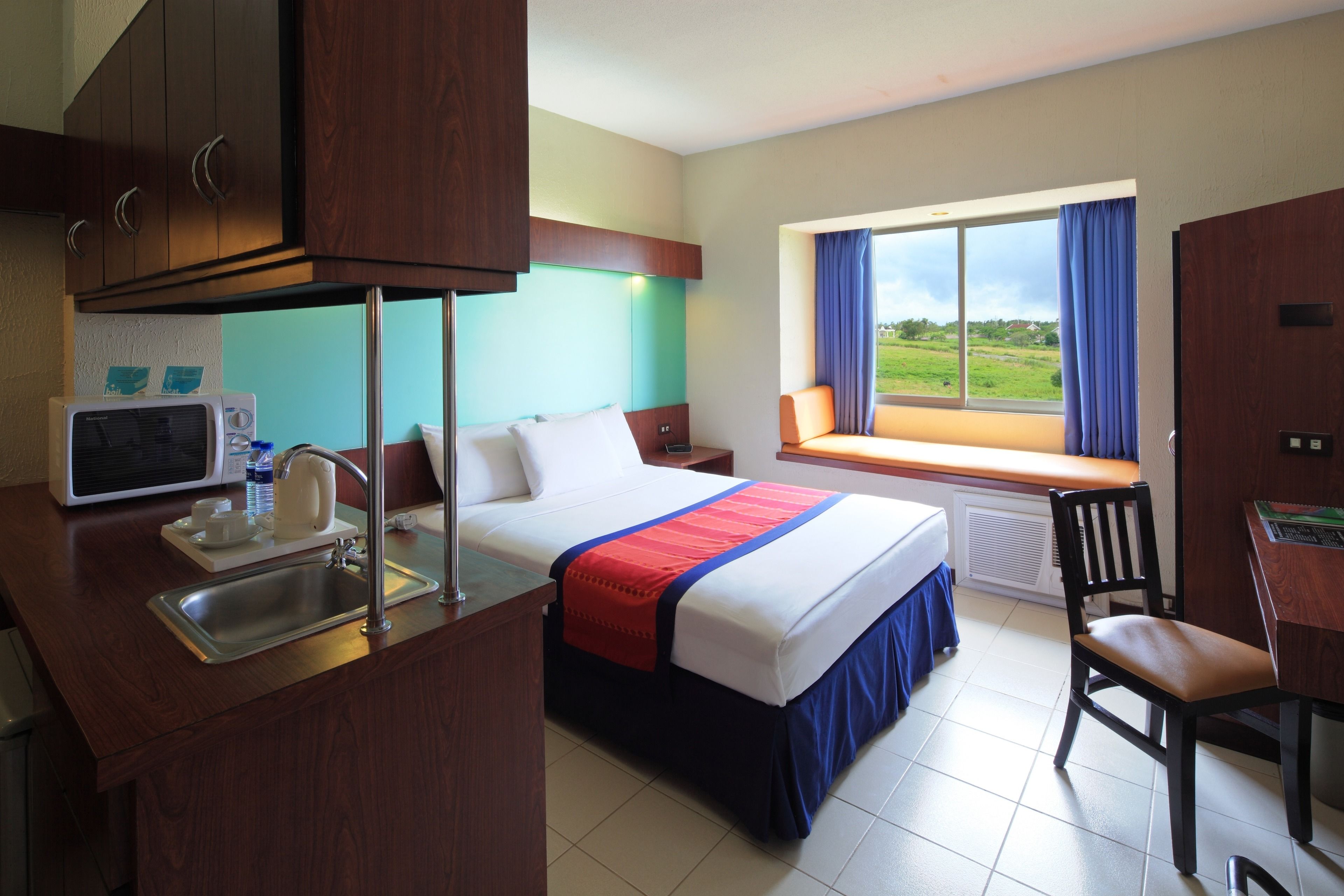 A hotel room with a bed, a desk with a chair, a microwave, a sink, and a window with a view of the outdoors.