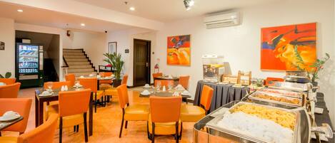 A dining area with orange chairs, tables set for meals, buffet with various food options, and a staircase in the background.