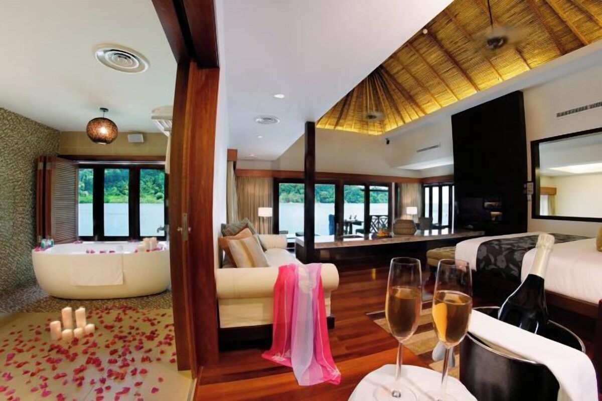A room with a large window, a jacuzzi, a sofa, a dining table with glasses and a bottle, and a television.