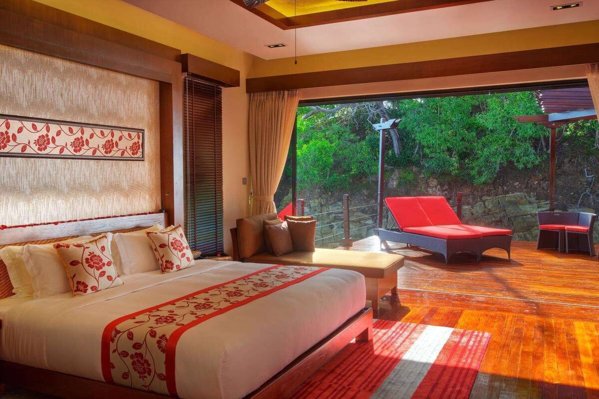 A bedroom with a large bed, a sofa, and a red chair. There is a view of greenery outside the window.