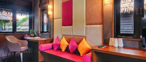 A hotel reception area with a wooden reception desk, a cushioned bench with colorful pillows, a chair, and a view of the outside through large windows.