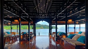 An open-plan lounge area with wooden flooring and seating, overlooking a body of water and greenery. - Gayana Marine Resort (Gaya Island)