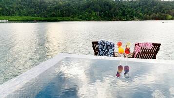 A poolside area with a table and two chairs, overlooking a calm lake and a forested hill.