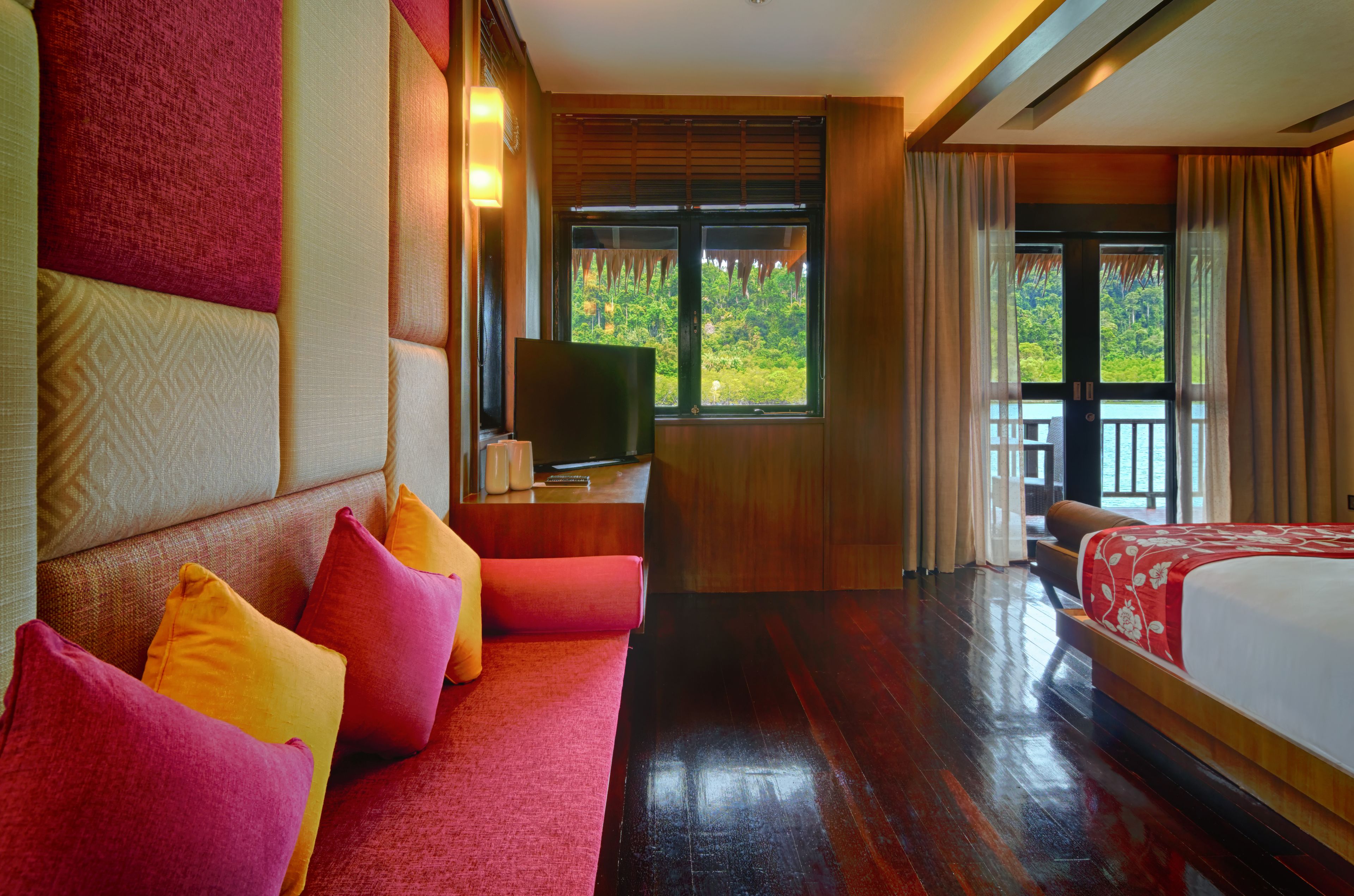 A hotel room with a large bed, a sofa with colorful cushions, a television, and a window with a view of greenery.