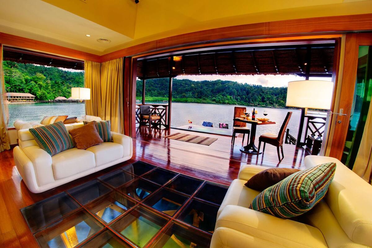 A spacious room with a large window overlooking a body of water and lush greenery. There is a white sofa with striped cushions and a dining area with a table and chairs.