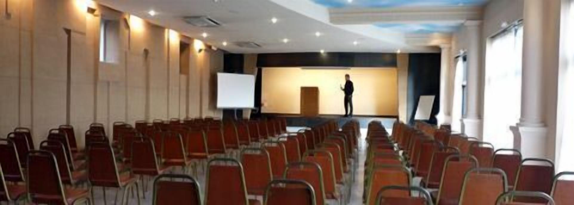 Un salón de conferencias con filas de sillas orientadas hacia un escenario donde hay una persona de pie.