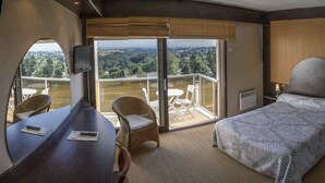 Quarto de hotel com cama, escrivaninha, duas cadeiras, televisão e varanda com vista para o campo.