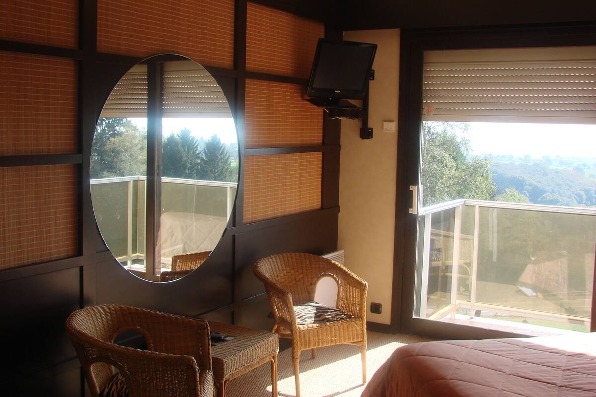A hotel room with a large mirror, two wicker chairs, a bed, and a balcony with a view of the landscape.