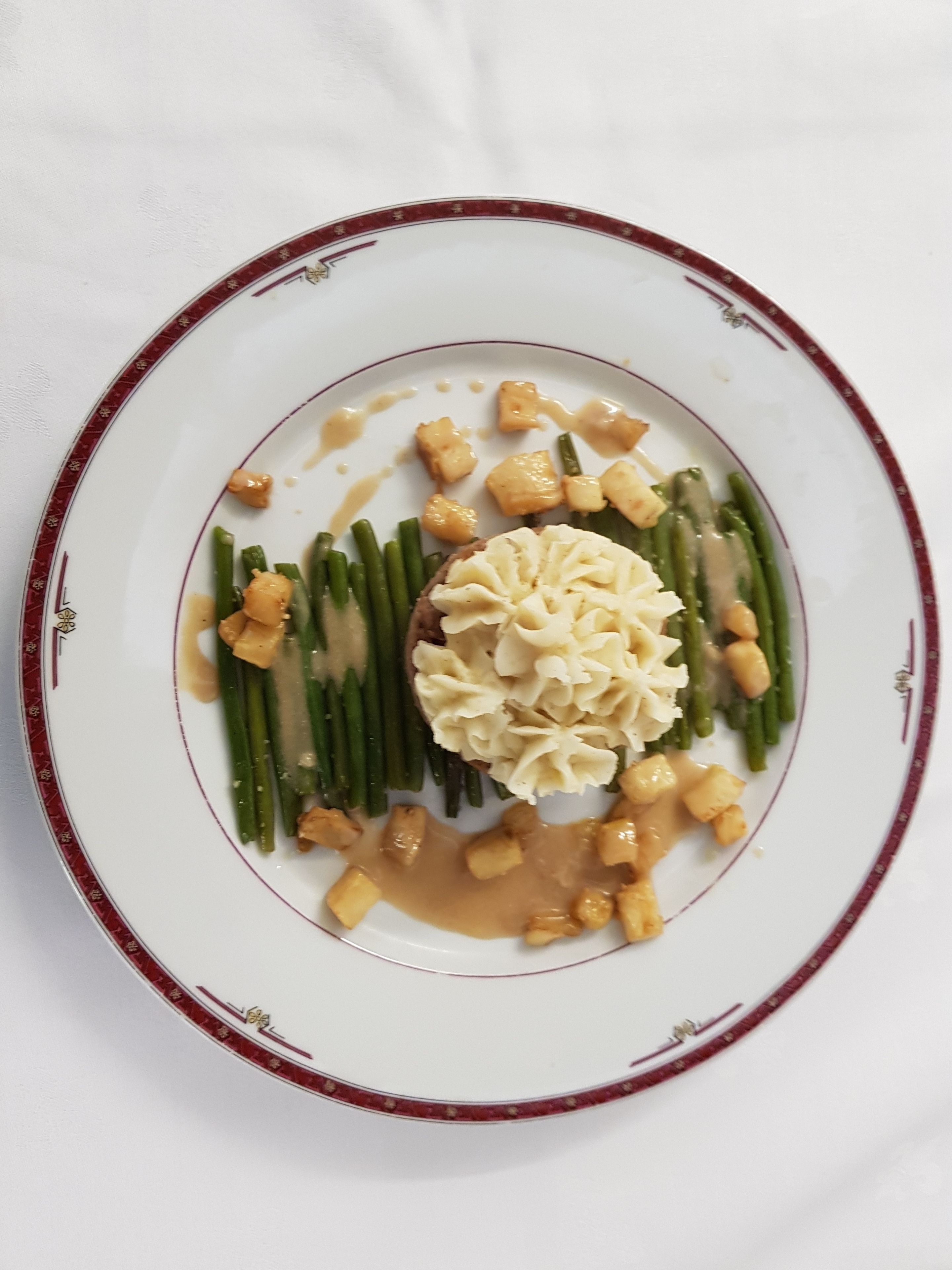 Ein Teller mit weißem Grund und rotem Rand, belegt mit grünen Bohnen, Kartoffelstampf und einer zentralen Häufung, möglicherweise ein Fleisch- oder Gemüsespezial, mit cremiger Sauce.