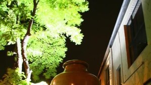 Un grand vase en céramique, placé sur un socle dans un espace extérieur, avec un arbre et un bâtiment en arrière-plan.