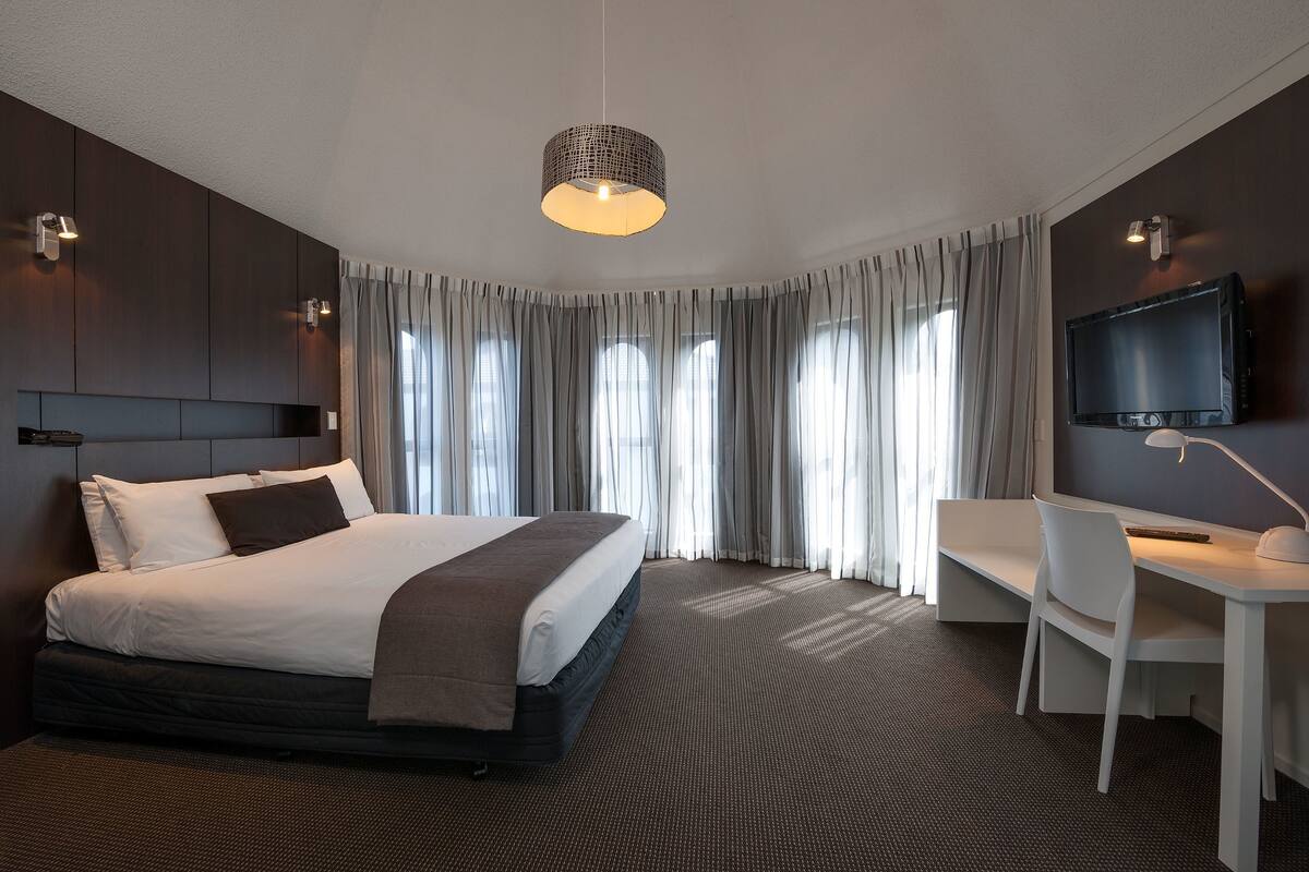 Une chambre d’hôtel moderne dotée d’un grand lit, d’un bureau, d’une télévision et d’une grande fenêtre avec des rideaux.