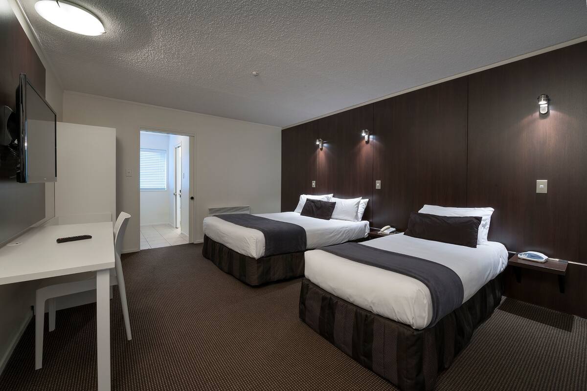 Une chambre d’hôtel avec deux lits, un bureau et une chaise. Il y a une porte qui donne sur une autre pièce et une fenêtre avec des stores.
