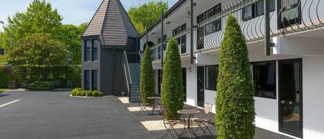 Un edificio moderno con tetto triangolare, un balcone con ringhiere in metallo e un cortile lastricato con tavoli e sedie.