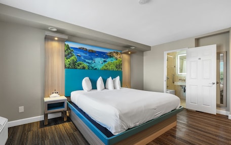 A modern bedroom with a large bed, a nightstand, a painting of a coastal scene, and a bathroom visible through an open door.