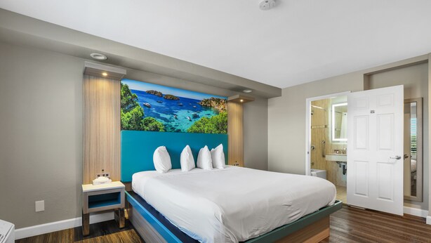 A modern bedroom with a large bed, a nightstand, a painting of a coastal scene, and a bathroom visible through an open door. - Motel Mediteran Escondido (Escondido)