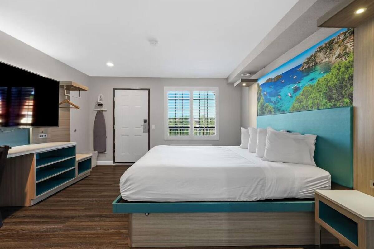A bedroom with a bed, a TV, a window, and a painting on the wall.