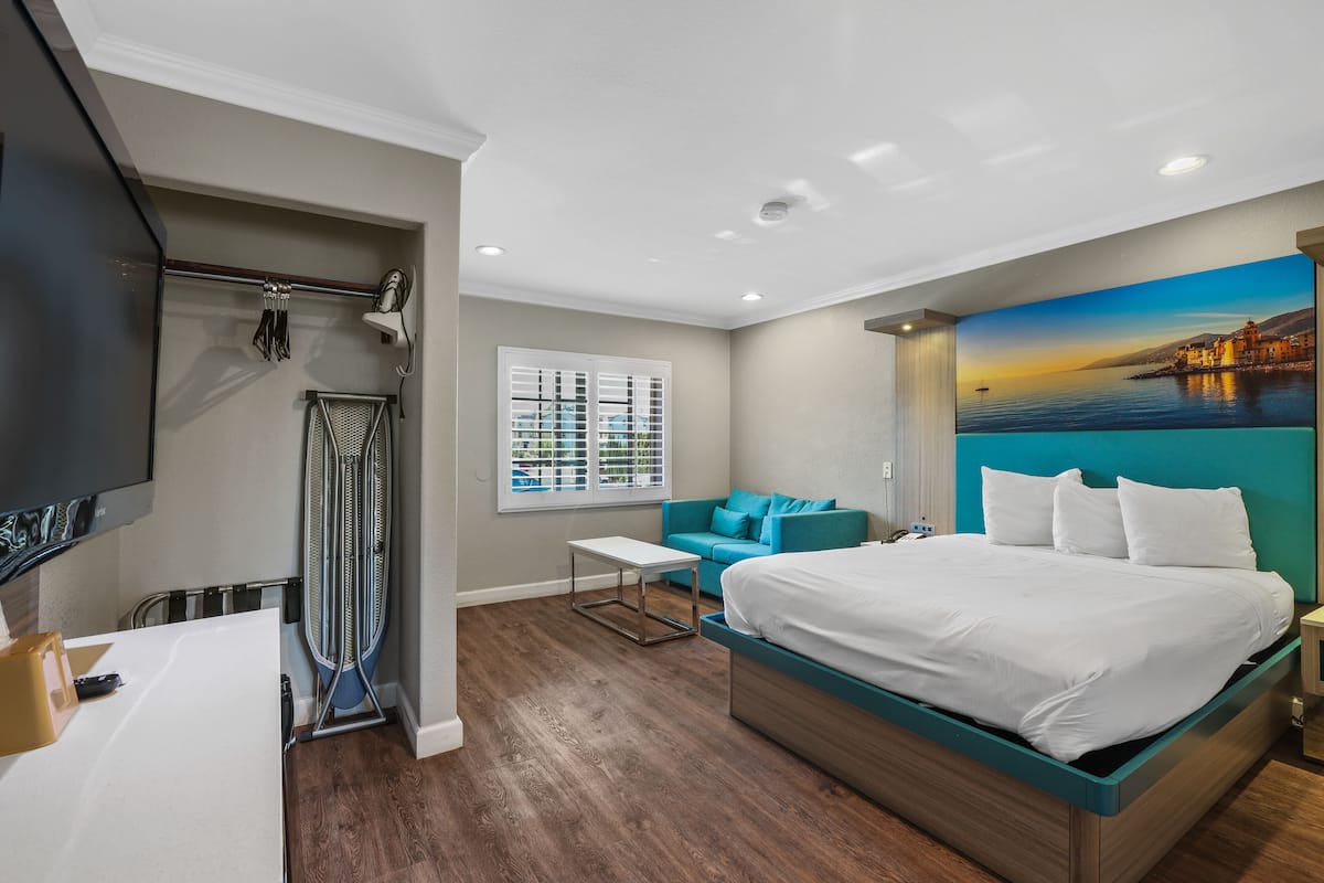 A modern hotel room with a large bed, a seating area with a sofa, a flat-screen TV, and a painting of a coastal scene.