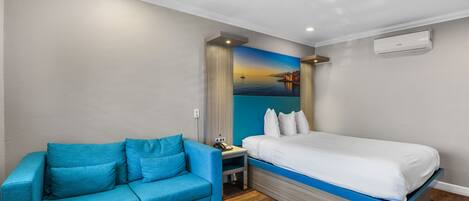 Una camera d'albergo moderna con un letto, un divano blu, un tavolino e un dipinto raffigurante un tramonto sull'acqua.
