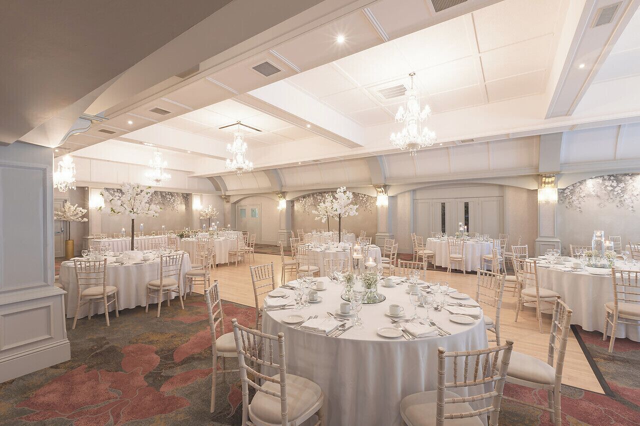 Une salle de banquet avec des tables rondes dressées pour le repas, des lustres et des compositions florales en guise de décoration centrale.