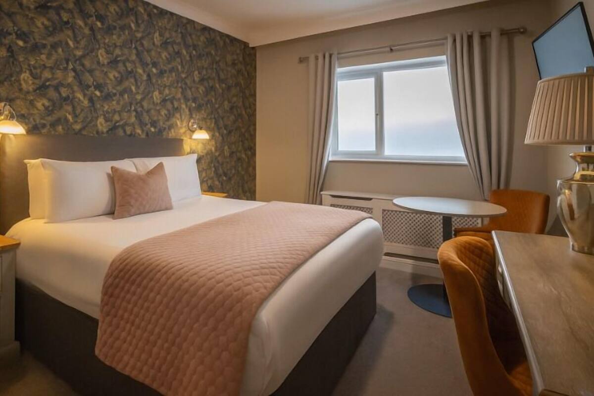 Quarto de hotel com cama, uma mesinha, uma cadeira, abajur e janela com cortinas.