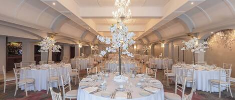 A banquet hall with round tables set for dining, white chairs, and a central chandelier.