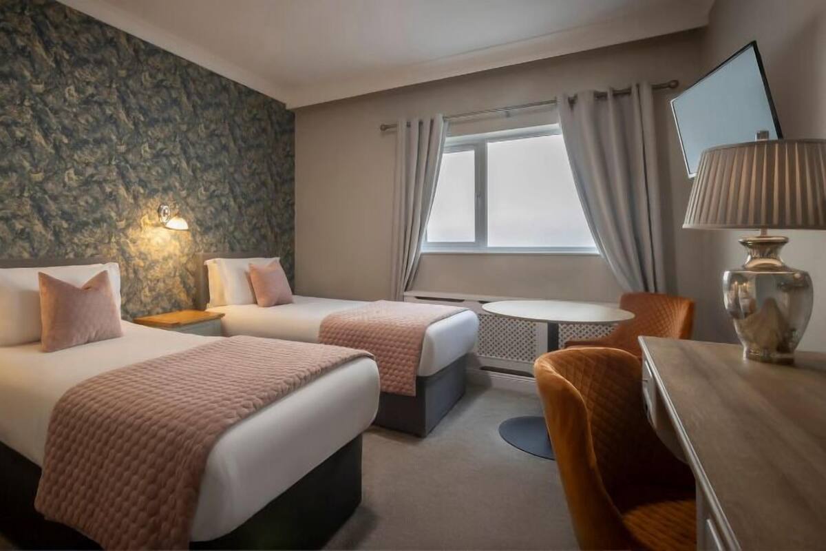 Quarto de hotel com duas camas, uma mesinha, uma cadeira, uma televisão e uma parede de pedra.