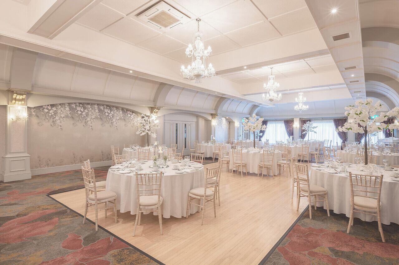 A spacious banquet hall with round tables, white tablecloths, and elegant chandeliers.