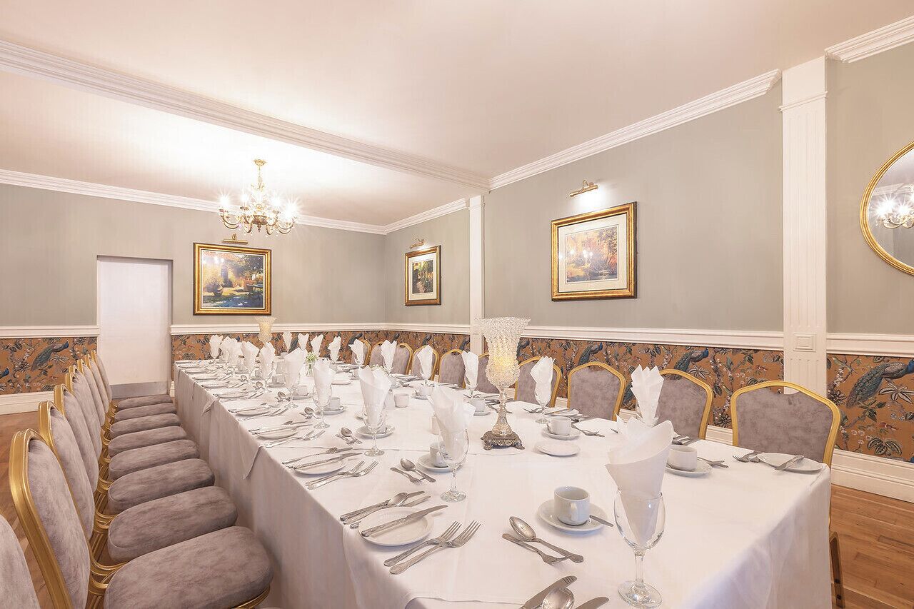 Un comedor formal con una larga mesa dispuesta para un banquete, flanqueada por filas de sillas tapizadas, y decorado con cuadros enmarcados y una araña.