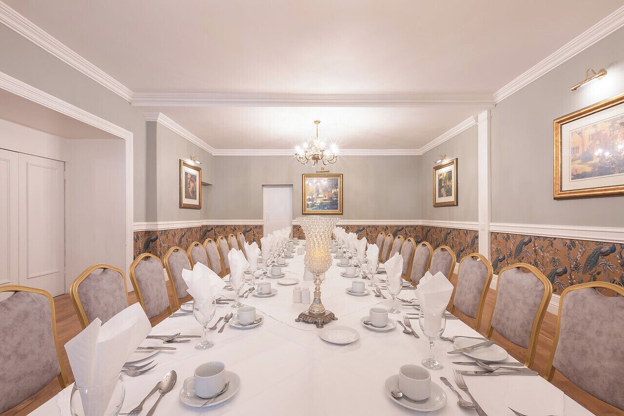 Une salle à manger élégante, avec une longue table dressée pour un banquet, entourée de chaises rembourrées, d’un lustre et d’œuvres d’art encadrées accrochées aux murs.