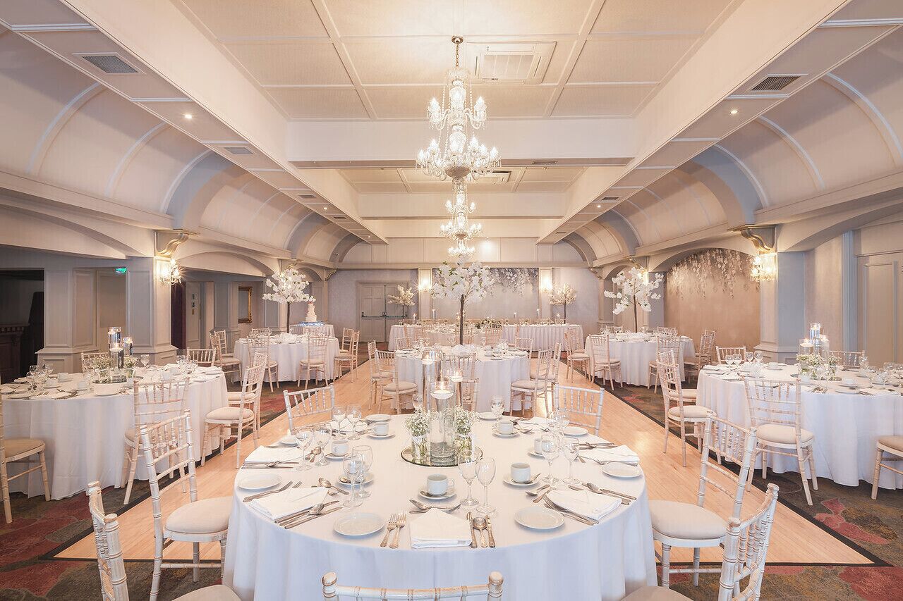 Un salón de banquetes con mesas redondas dispuestas para un evento formal, con candelabros y arreglos florales blancos.