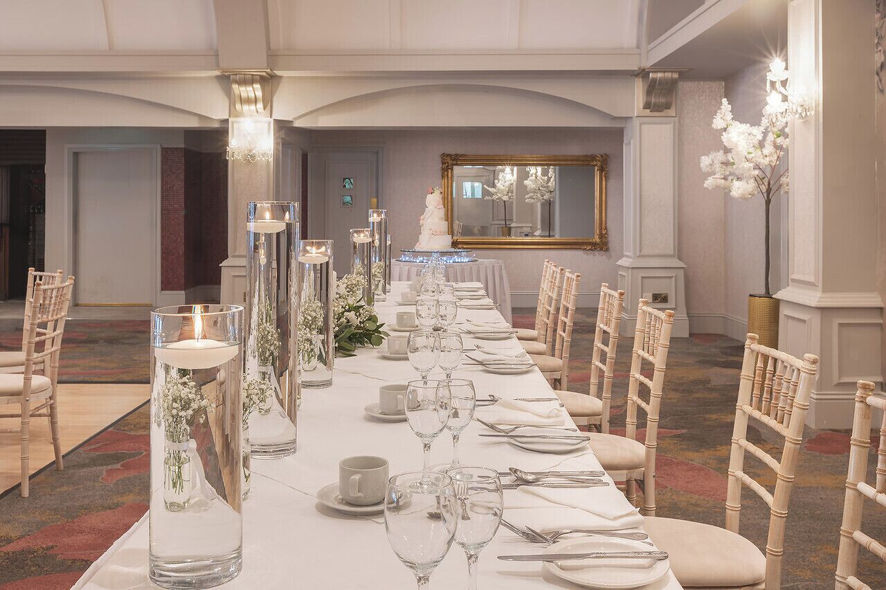 A long banquet table set with glassware, candles, and floral arrangements in a formal dining room with chandeliers and a mirror on the wall.
