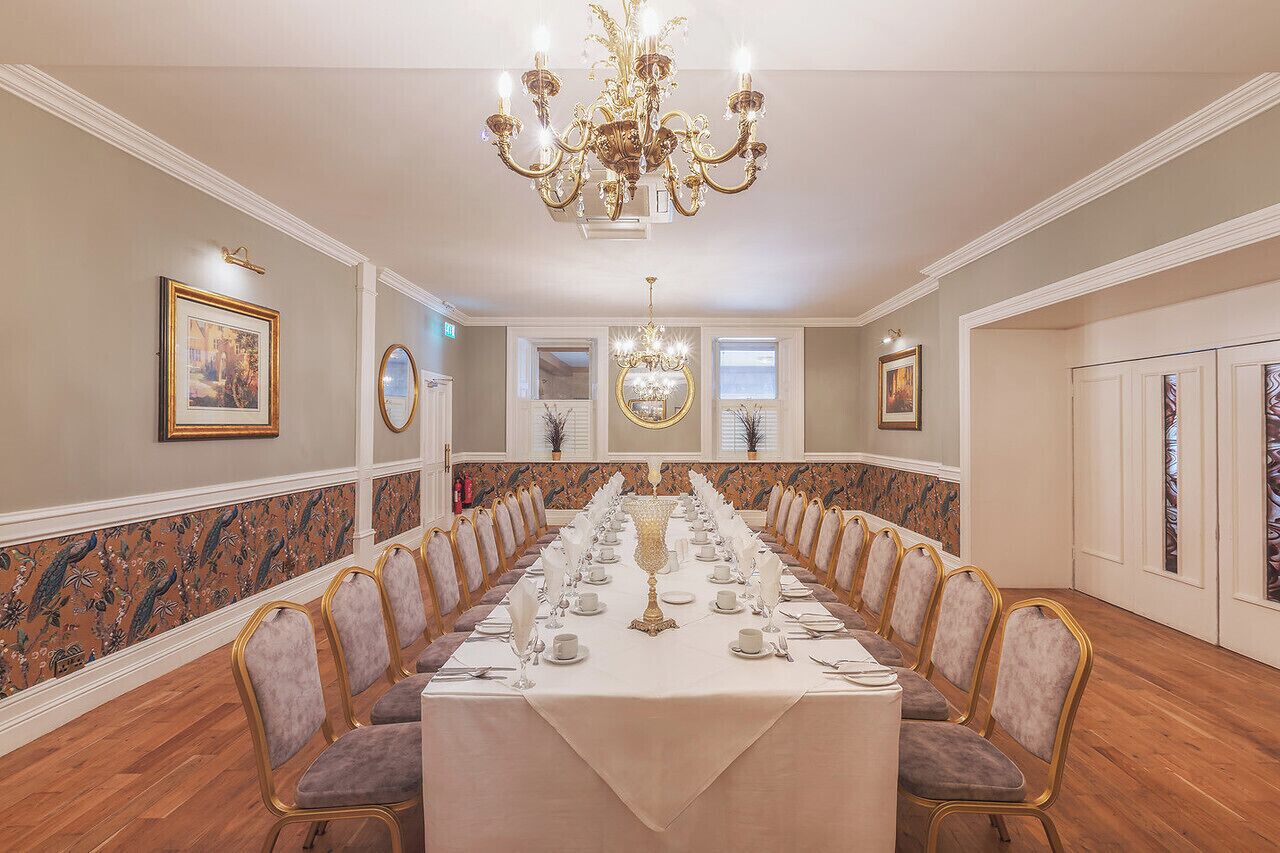 A formal dining room with a long table set for a banquet, surrounded by upholstered chairs, a chandelier, and framed artwork on the walls.