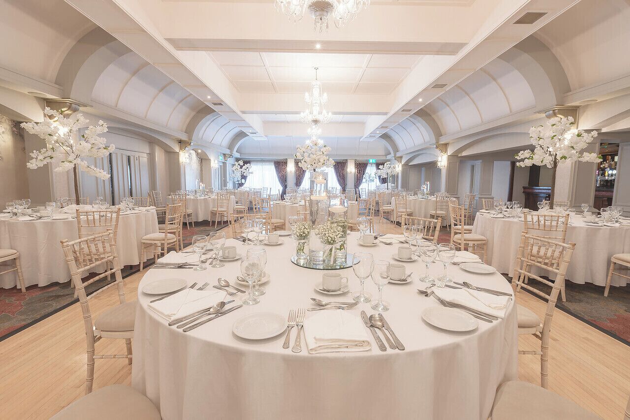 A banquet hall with round tables set for a formal event, featuring white tablecloths, floral centerpieces, and elegant lighting fixtures.