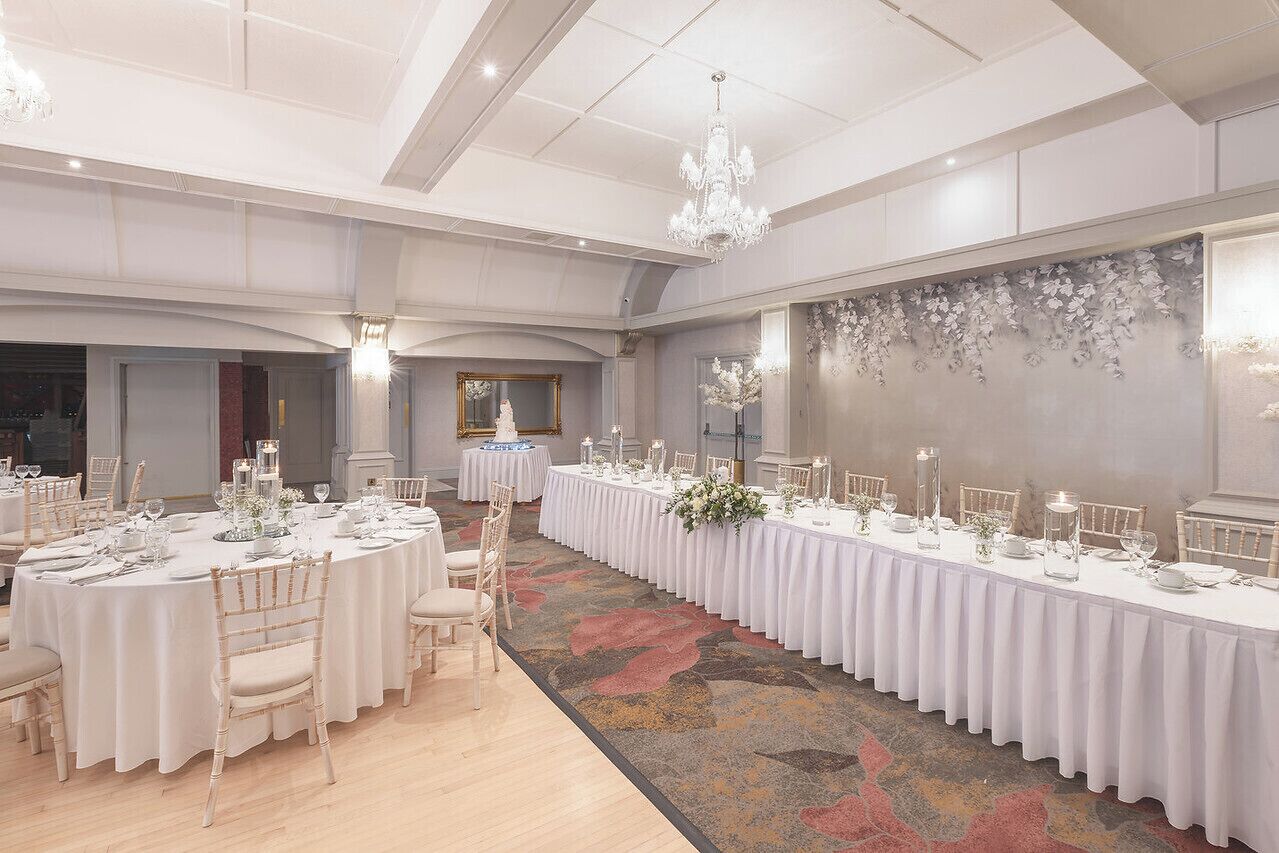 A banquet hall with round tables set for a formal event, featuring white tablecloths, floral centerpieces, and candle holders.