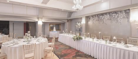Ein Festsaal mit runden Tischen, festlich gedeckt für eine Veranstaltung, mit weißen Tischdecken, floralen Mittelpunkten und Kerzenständern.