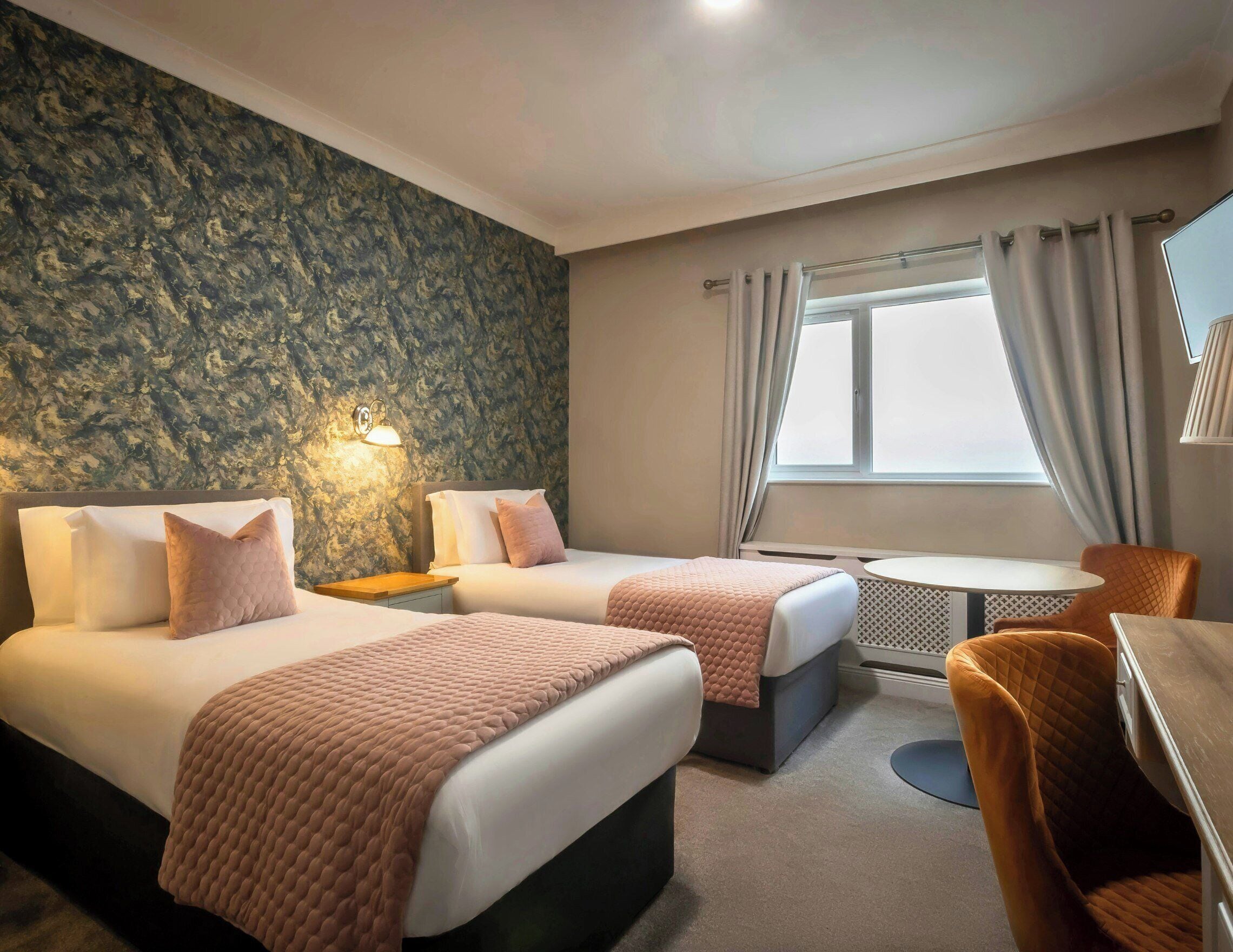 Ein Hotelzimmer mit zwei Betten, einem kleinen Tisch, einem Stuhl und einem Fenster mit Vorhängen.
