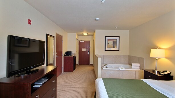 A hotel room with a flat-screen TV, a bed with a green bedspread, a bathroom with a bathtub, and a desk with a lamp. - Cobblestone Inn & Suites - Brillion (Brillion)