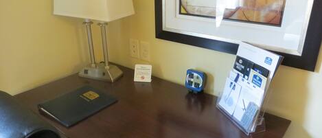 A hotel room desk with a lamp, a sign indicating a non-smoking room, a power outlet, and a framed picture on the wall.