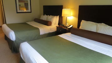 A hotel room with two beds, each with green bedding and white pillows. There is a nightstand with a lamp, a clock, and a phone. A framed picture hangs on the wall above the beds.