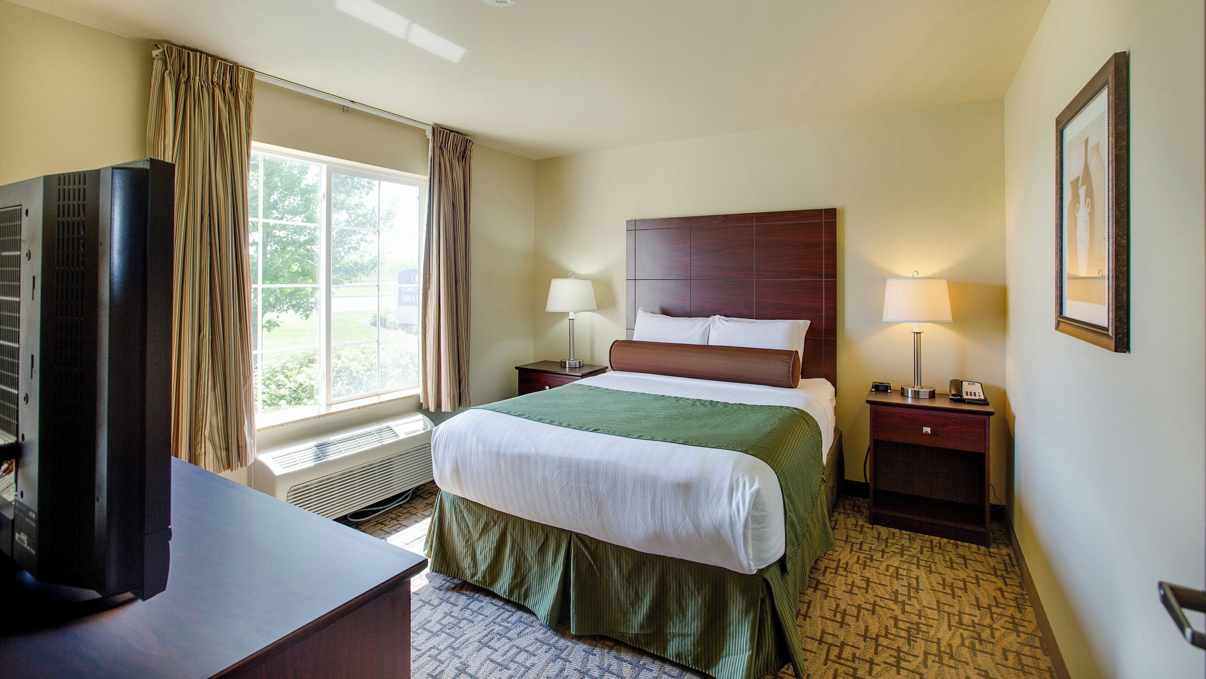 A hotel room with a bed, two bedside tables, a TV, a window with curtains, and a framed picture on the wall.