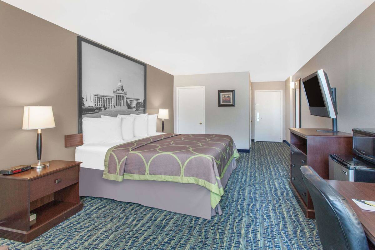 Ein Hotelzimmer mit einem Bett, einem Schreibtisch mit Fernseher, einer Lampe und einem gerahmten Bild an der Wand.