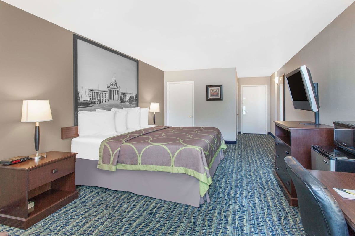 Ein Hotelzimmer mit einem Bett, einem Schreibtisch mit Fernseher, einer Lampe und einem gerahmten Bild an der Wand.