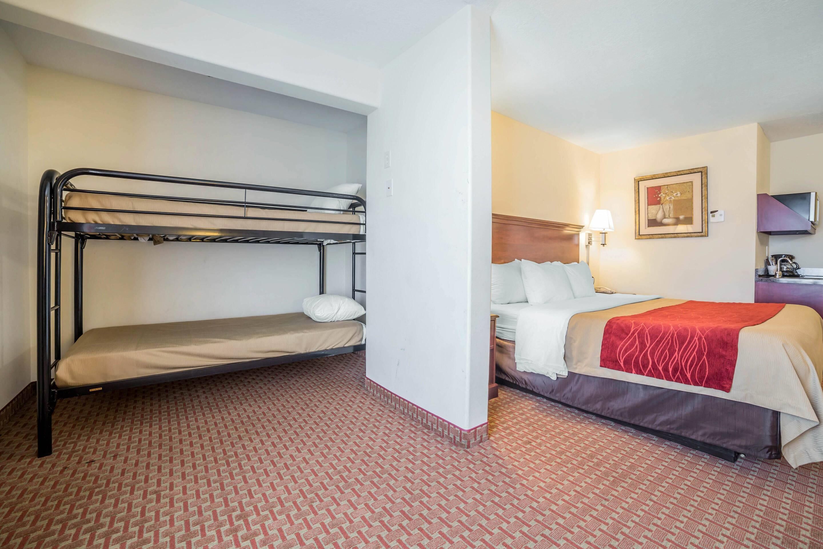 A hotel room with a bunk bed, a single bed, a bedside table with a lamp, and a framed picture on the wall.