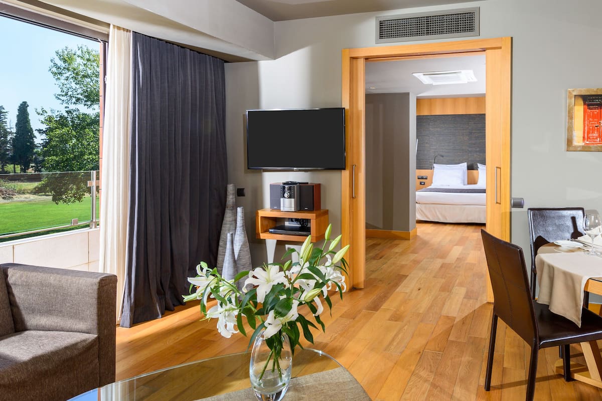 Une chambre d’hôtel moderne dotée d’une grande fenêtre, d’un téléviseur à écran plat, d’un coin repas avec une table et des chaises, et d’un lit avec une tête de lit en bois.