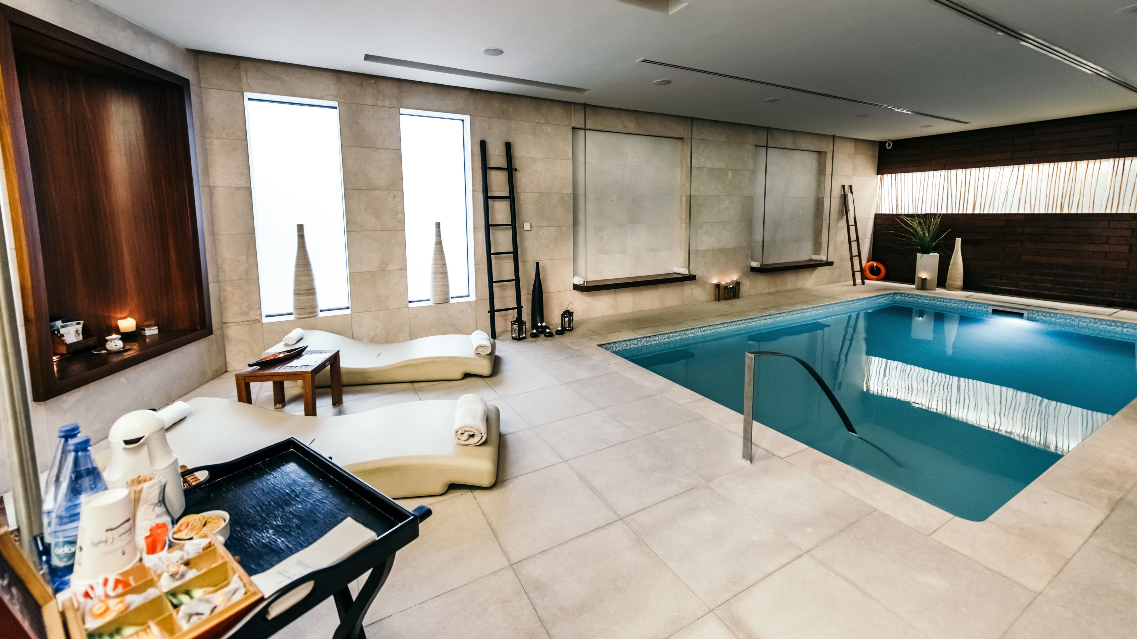 A modern indoor swimming pool with a lounge area, including a massage chair, a tray table with snacks, and a ladder for pool access.
