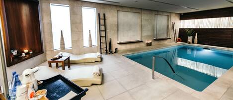 A modern indoor swimming pool with a lounge area, including a massage chair, a tray table with snacks, and a ladder for pool access.