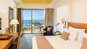 Hotelkamer met een bed, bureau met lamp, stoel, klein tafeltje met fles en glas, en uitzicht op zee door een glazen deur.