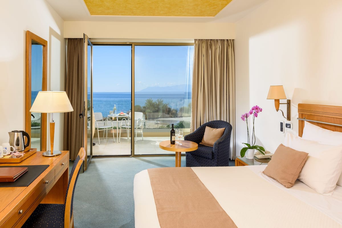 Une chambre d’hôtel avec un lit, un bureau doté d’une lampe, une chaise, une petite table avec une bouteille et un verre, et une vue sur la mer par une porte vitrée.